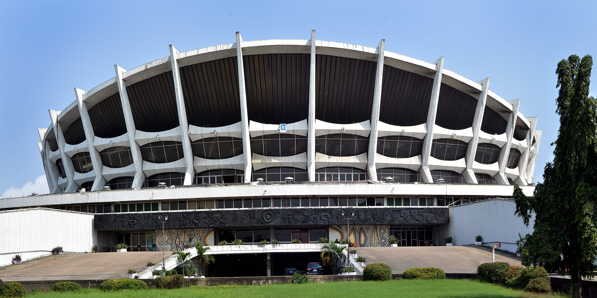 The white, ribbed, saucer-shaped roof of the National Theatre in Iganmu, Lagos, above a dark decorated facade, with a lawn and trees in front under a blue sky