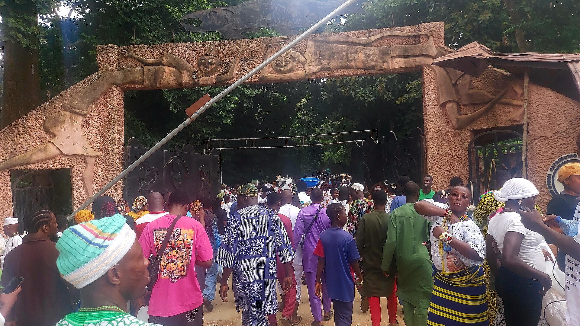 A crowd in colourful clothes walks through a stone gateway decorated with sculpted figures at the entrance to the Osun-Osogbo Sacred Grove, with dense forest behind