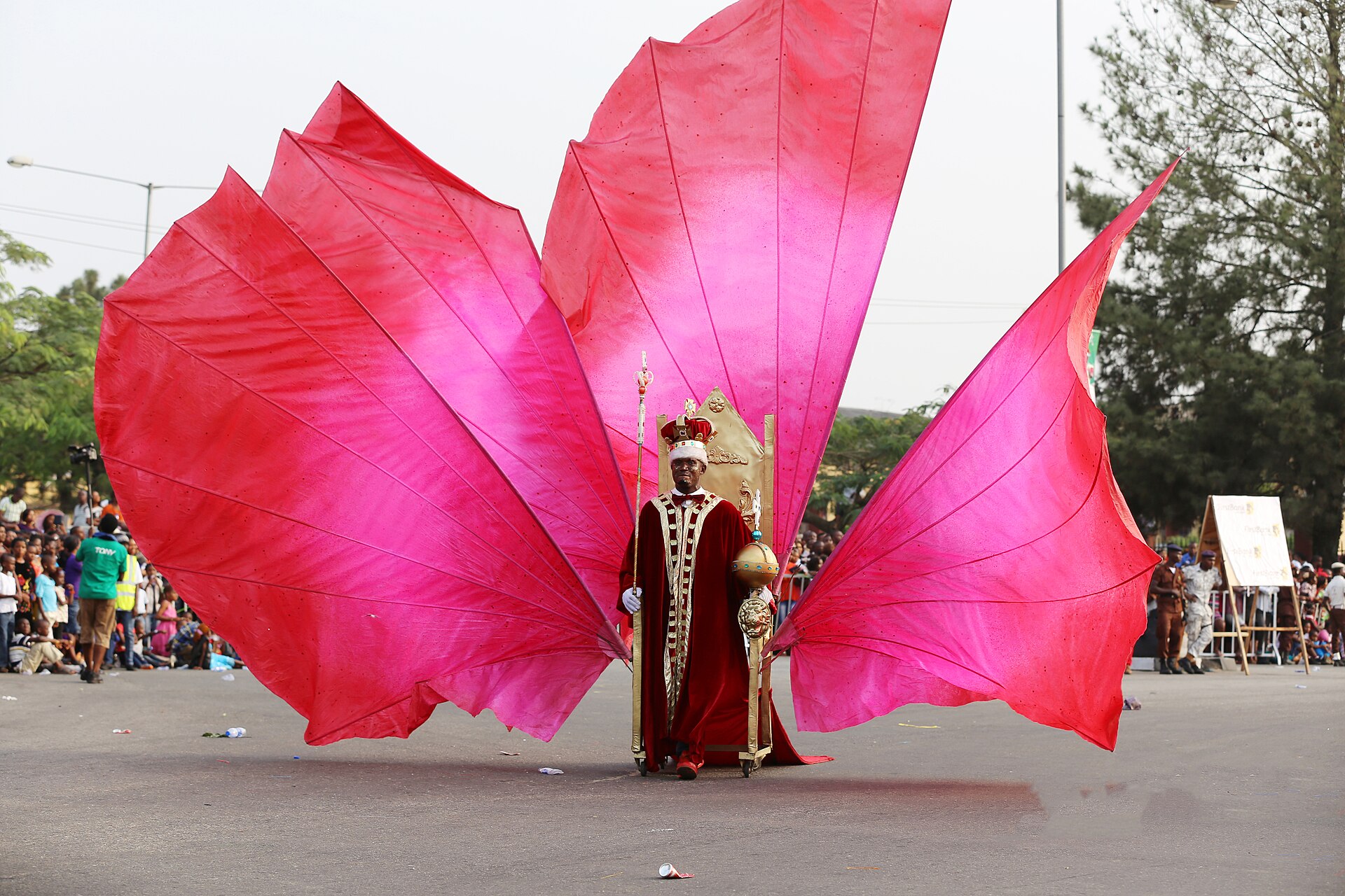 A performer in a red robe and crown holding a sceptre stands before a golden throne framed by giant pink fabric wings on a road lined with spectators