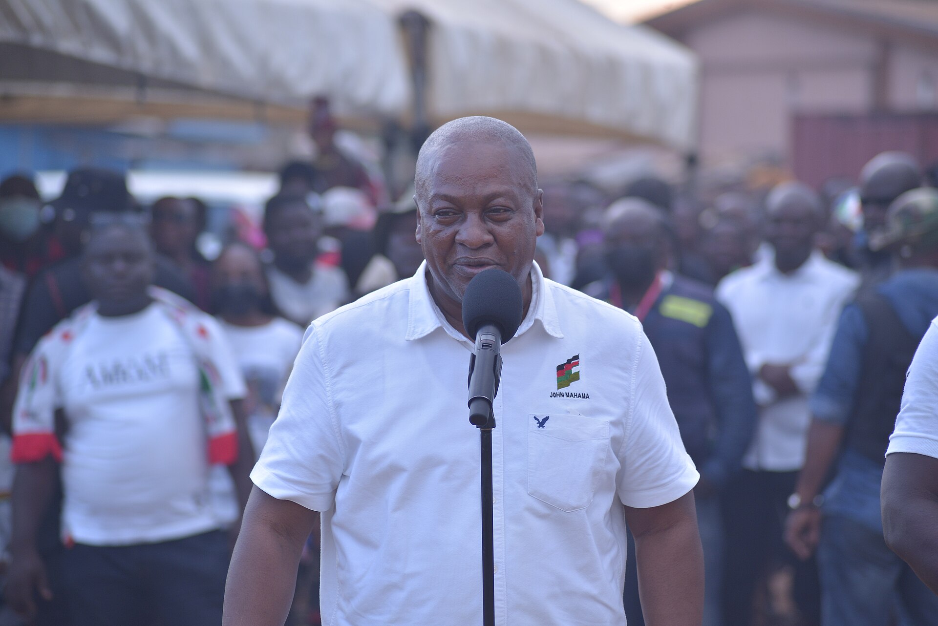 John Mahama in a white short-sleeved shirt speaks into a standing microphone in front of a crowd outdoors