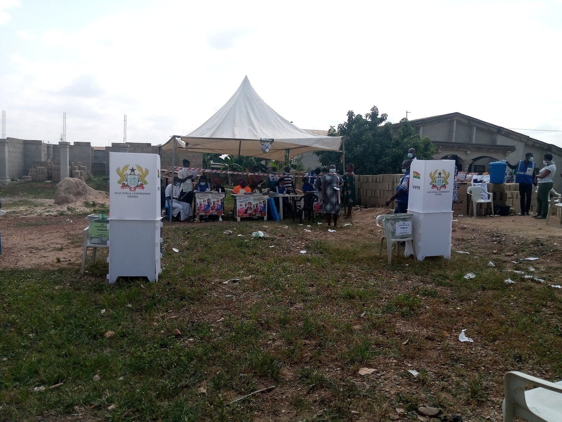 Two white Electoral Commission of Ghana voting screens stand on grass in front of a tent where poll officials and voters sit at tables