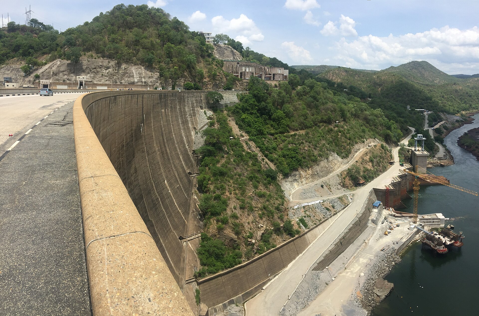 The curved concrete wall of the Kariba Dam seen from the road along its crest, with green hills and the Zambezi River gorge below