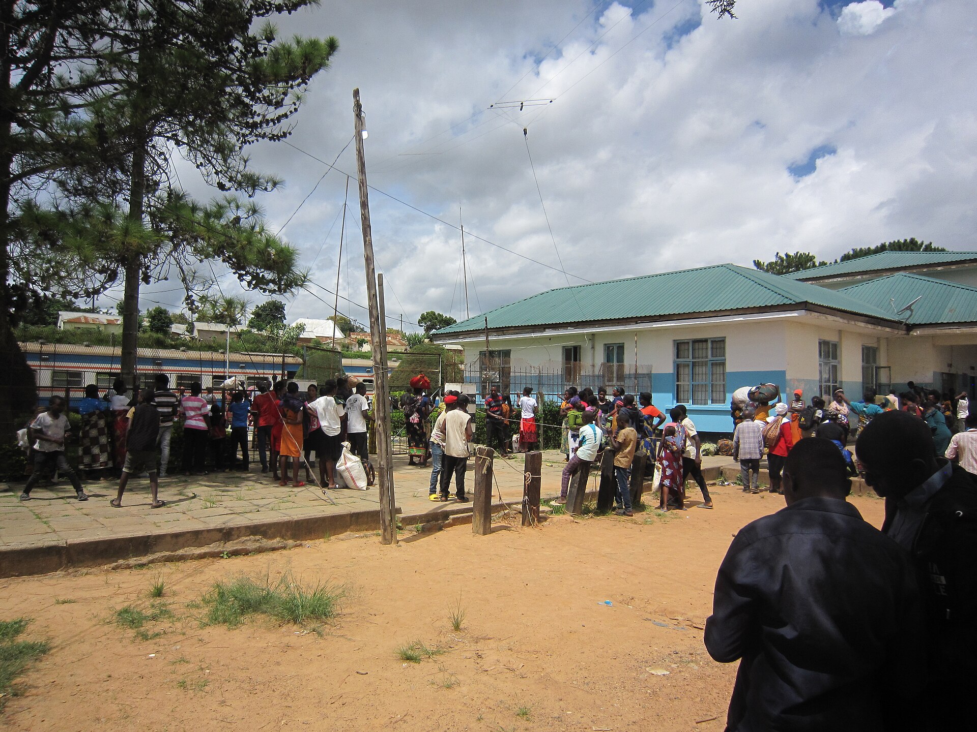 A crowd with bags and bundles waits outside a single-storey station building with a green roof, a blue-and-white TAZARA train behind them