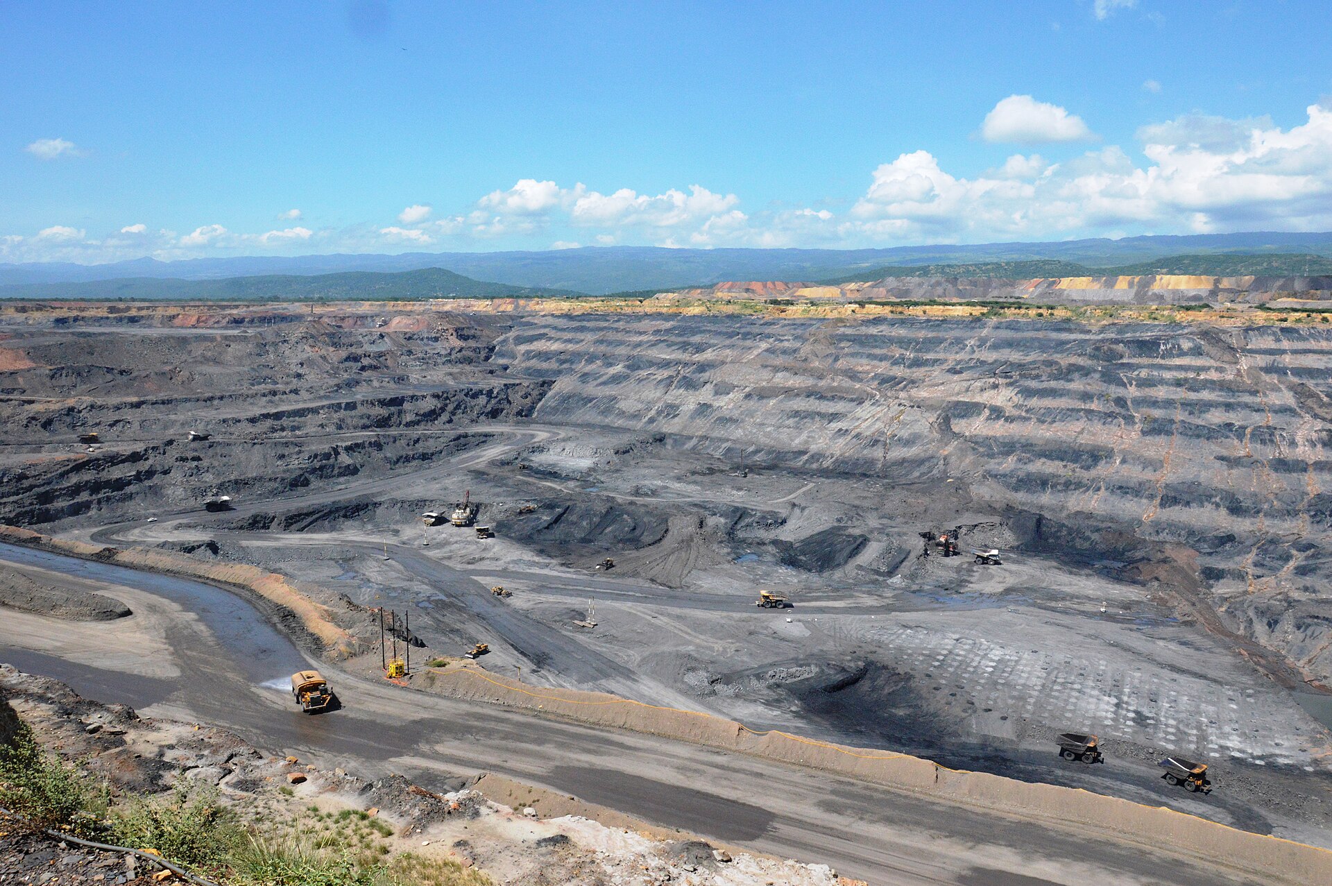 Open-pit Cerrejón coal mine in La Guajira with haul trucks on terraced grey benches under a blue sky with white clouds