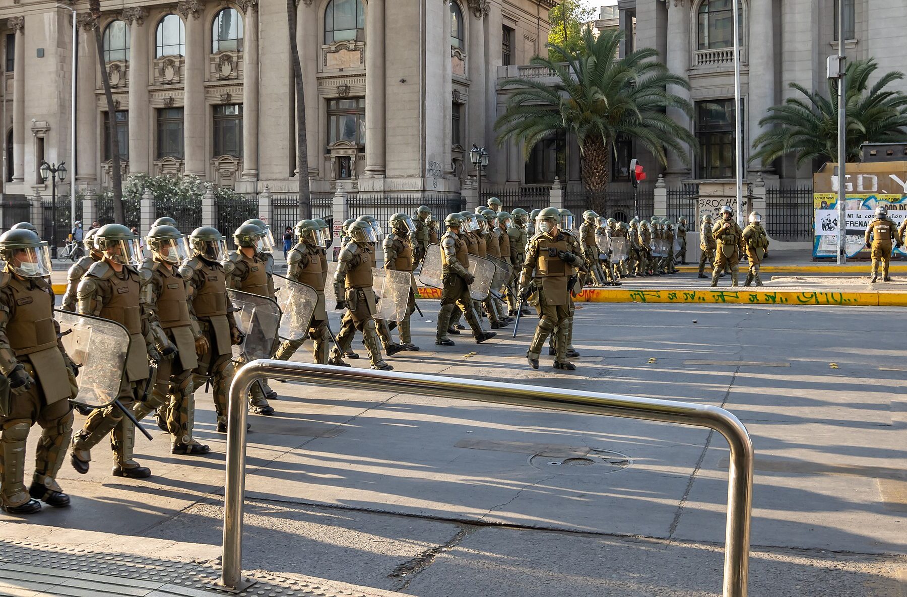 Carabineros officers in olive-green riot gear with helmets and shields walk along a city street past neoclassical buildings and palm trees