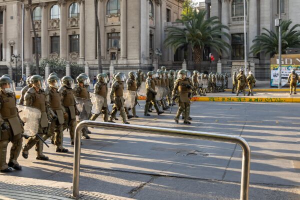 Carabineros officers in olive-green riot gear with helmets and shields walk along a city street past neoclassical buildings and palm trees