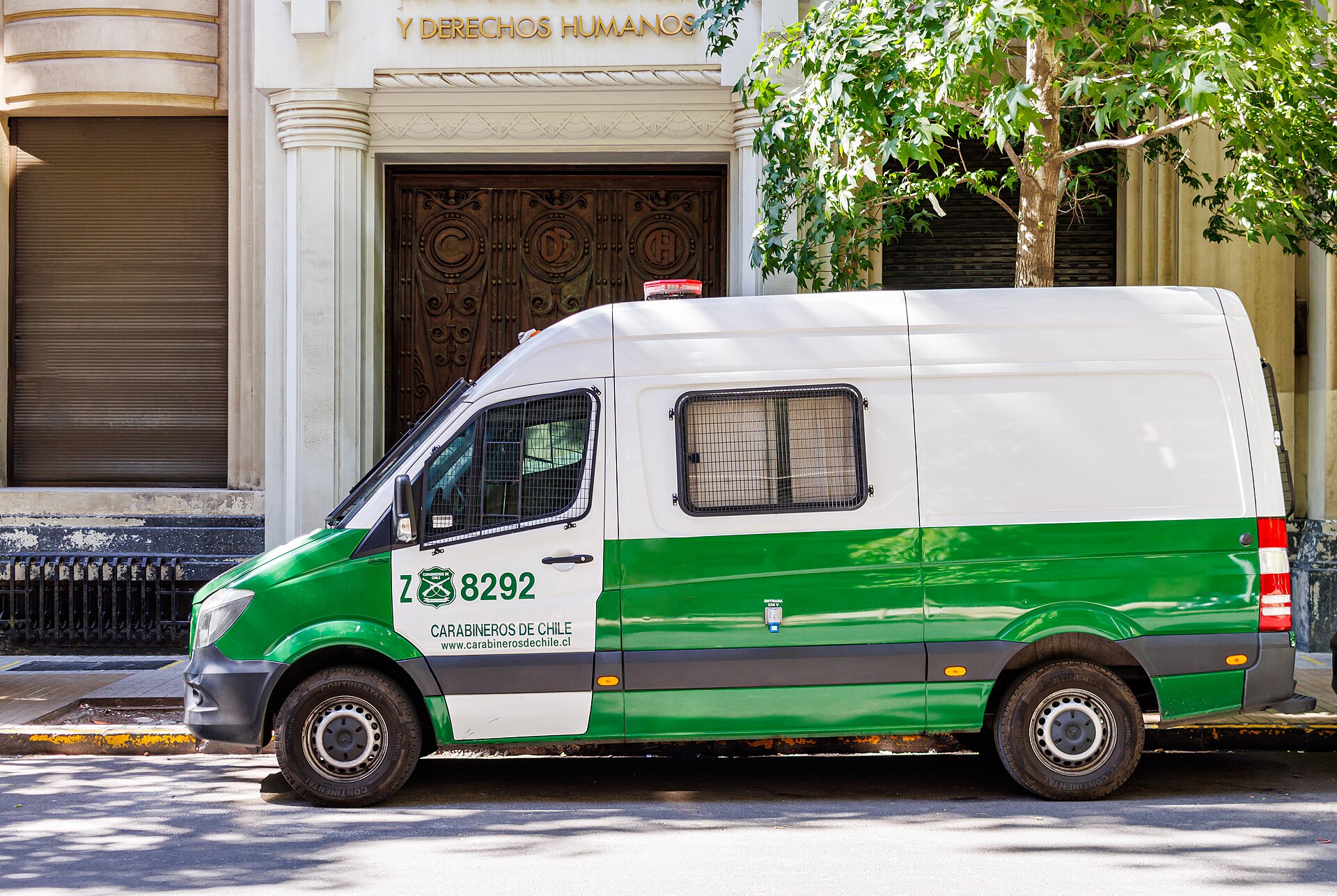 A green and white Carabineros de Chile police van with barred windows parked on a street in front of a stone building with carved wooden doors