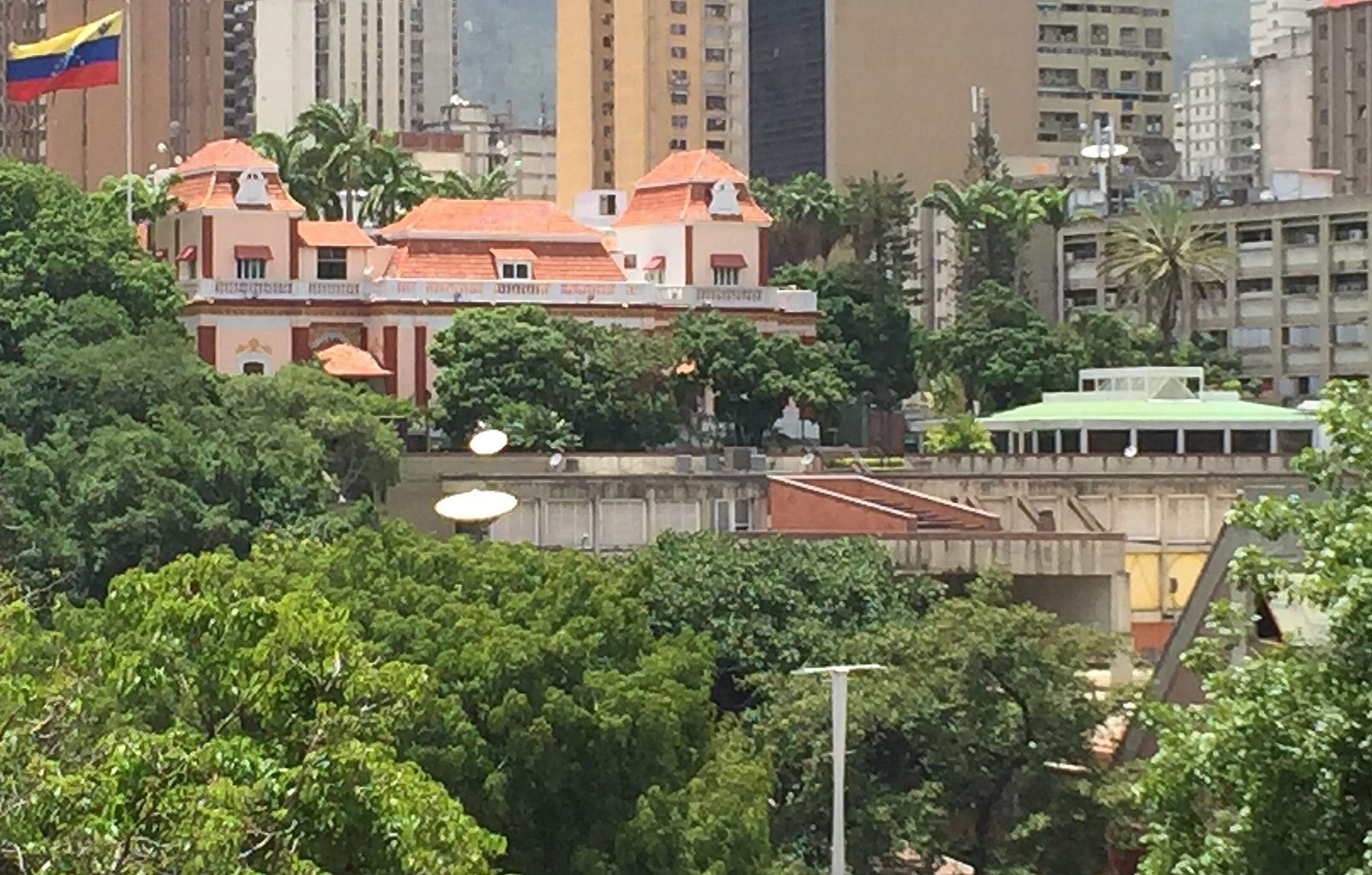 The red-roofed Miraflores Palace in Caracas among trees, with a Venezuelan flag flying at the left and office towers behind