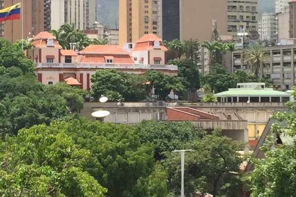 The red-roofed Miraflores Palace in Caracas among trees, with a Venezuelan flag flying at the left and office towers behind