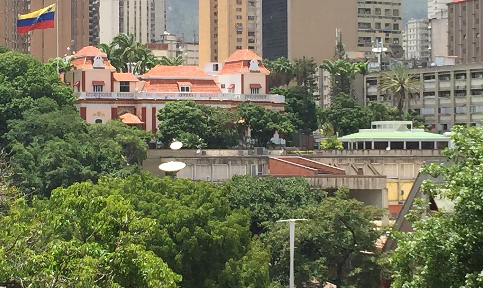 The red-roofed Miraflores Palace in Caracas among trees, with a Venezuelan flag flying at the left and office towers behind