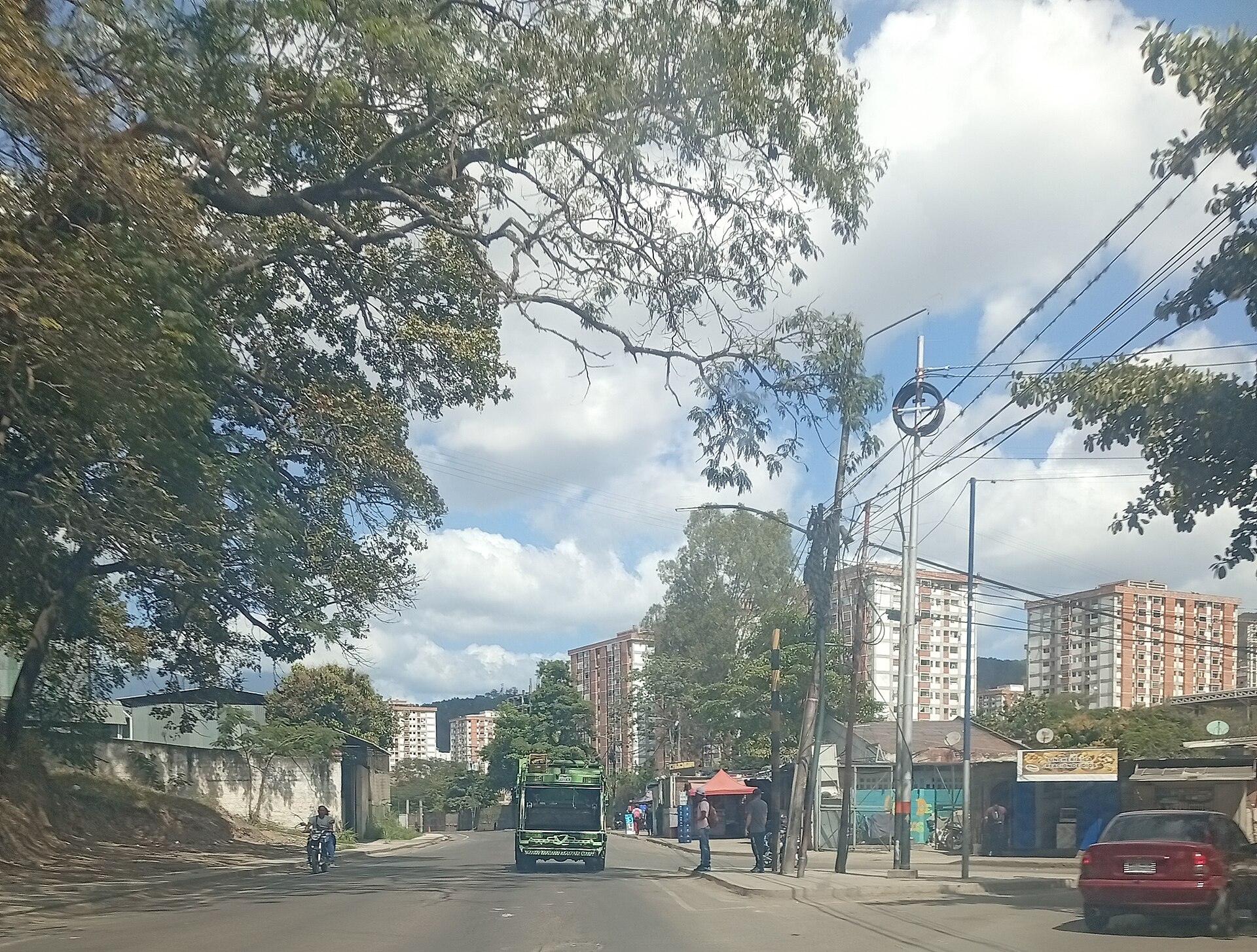 A Caracas street with a green truck, a motorcycle, roadside stalls and apartment towers behind trees under a cloudy sky