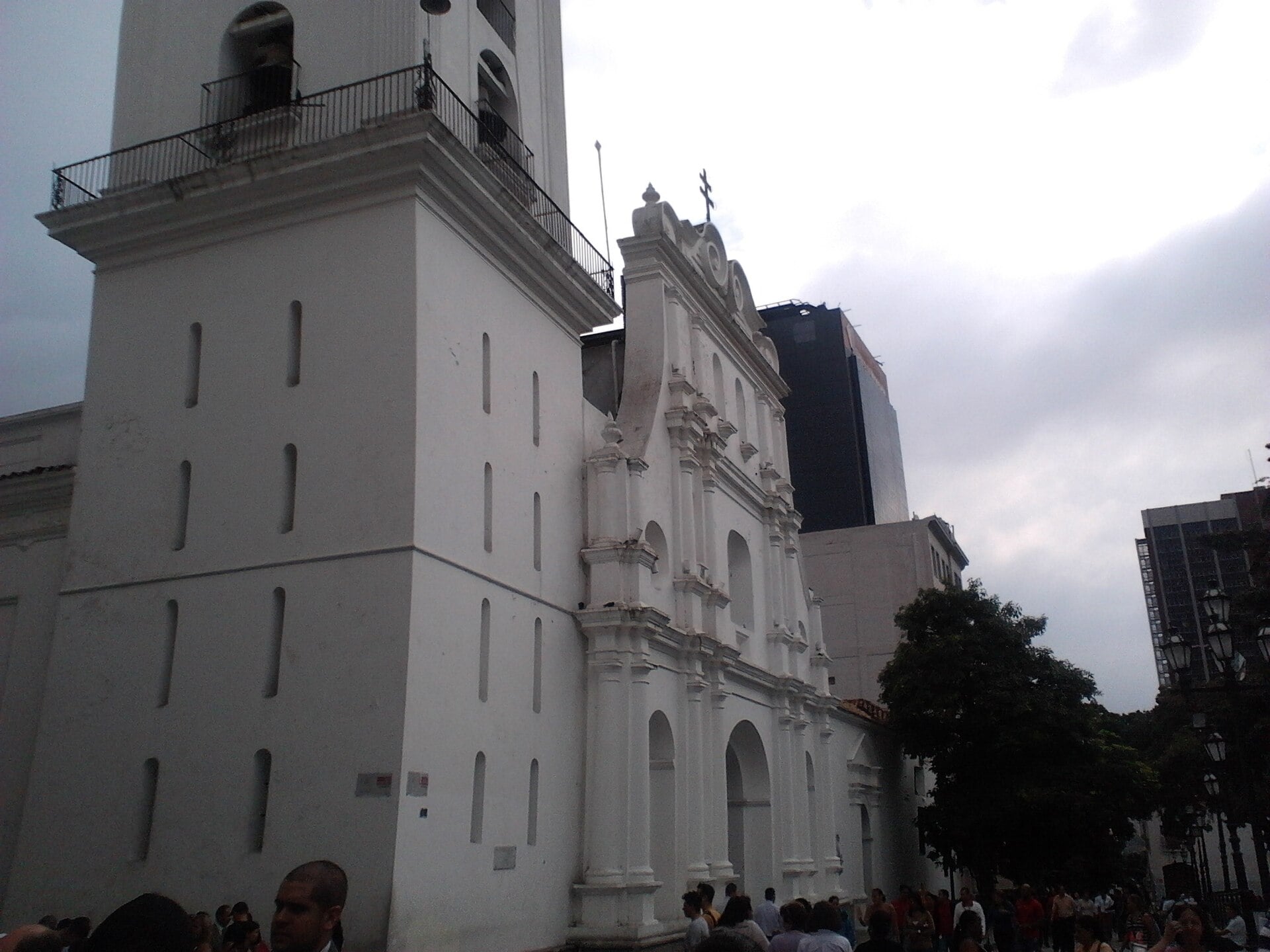 The white bell tower and baroque front of the Caracas Cathedral on a cloudy day, with people standing in the plaza in the foreground and office towers behind