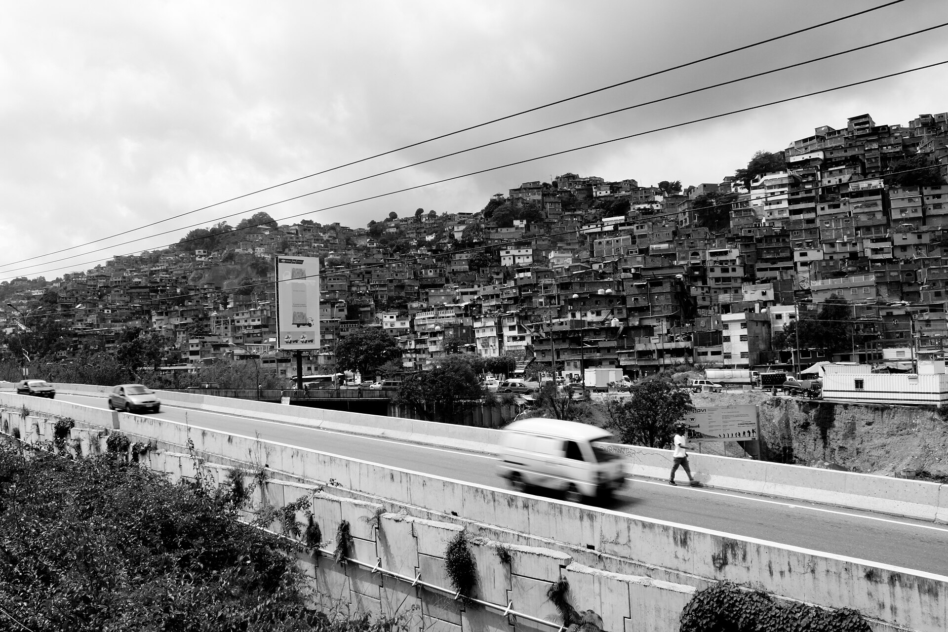 Black-and-white view of a steep hillside covered with dense brick homes in Petare above a highway with a white van and cars