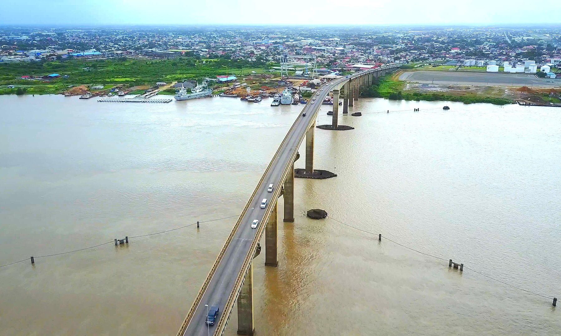 Aerial view of a long concrete road bridge with cars crossing a wide brown river, with a town, a quay and moored vessels on the left bank under a hazy sky