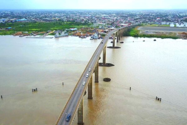 Aerial view of a long concrete road bridge with cars crossing a wide brown river, with a town, a quay and moored vessels on the left bank under a hazy sky