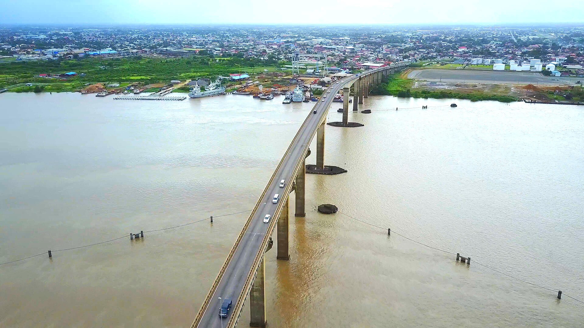 Aerial view of a long concrete road bridge with cars crossing a wide brown river, with a town, a quay and moored vessels on the left bank under a hazy sky