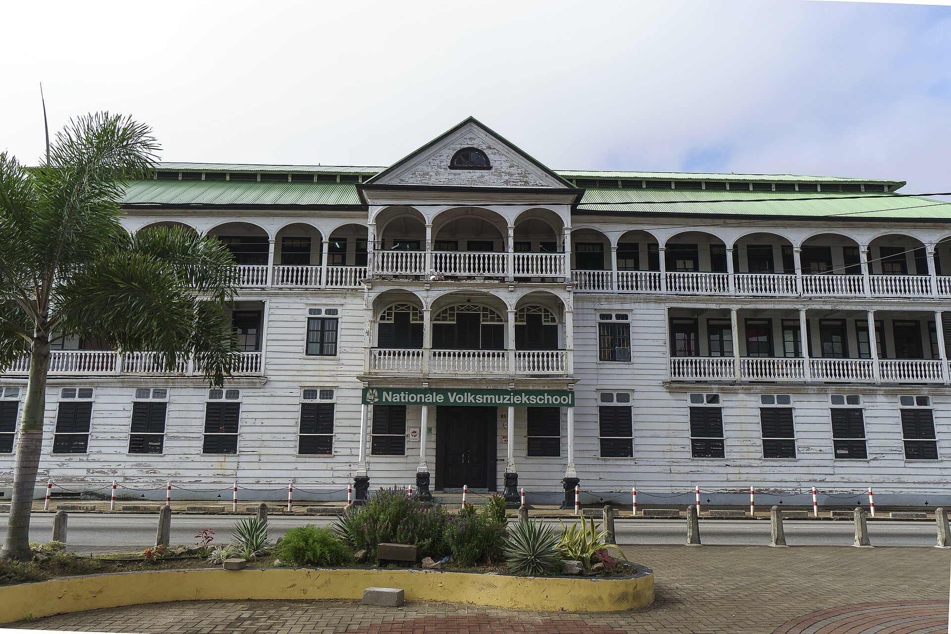 A white three-storey colonial wooden building with a green roof, arched verandas and a central gable, with a green sign reading Nationale Volksmuziekschool, seen across a street and a small garden