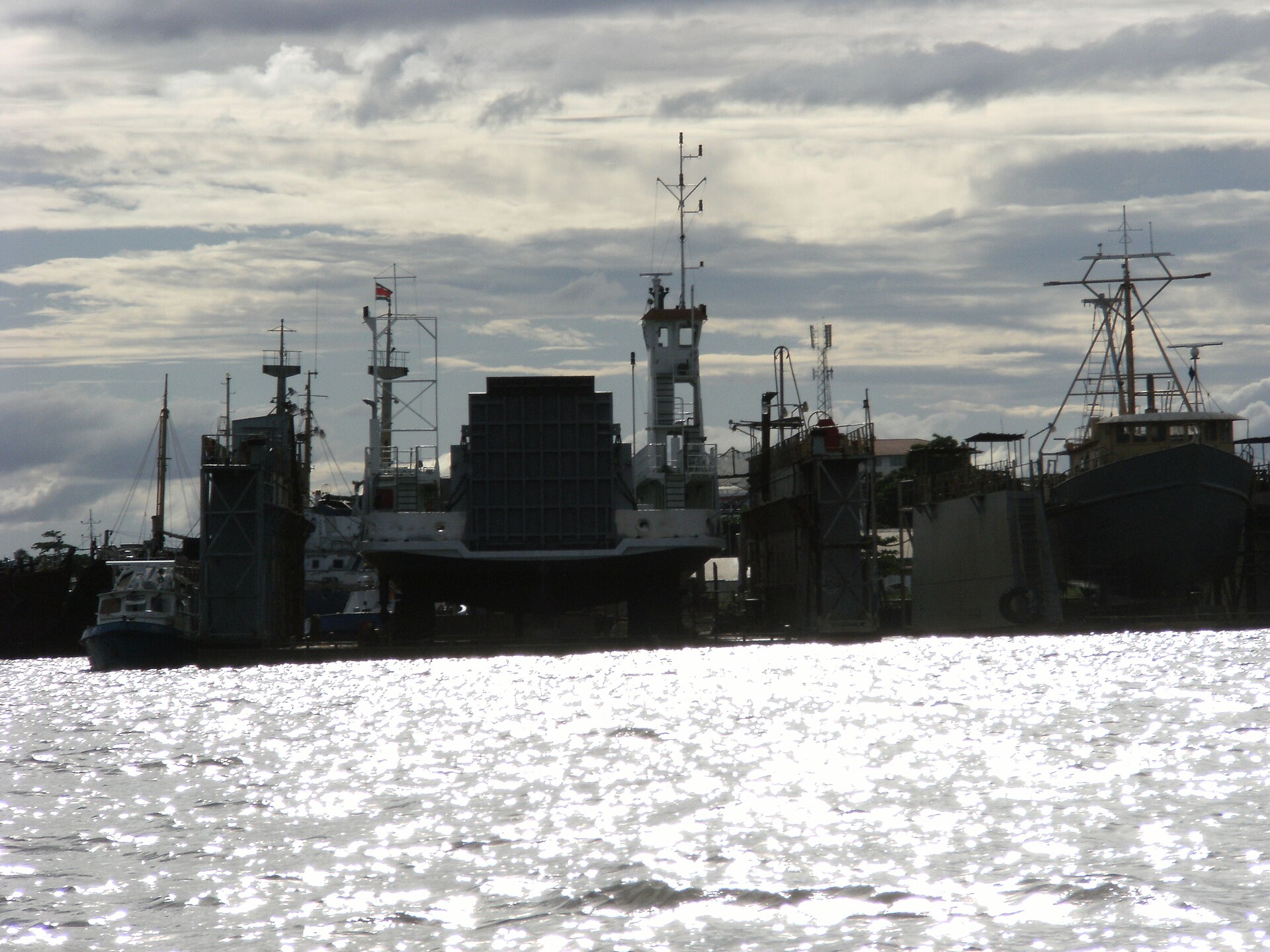 Backlit view across glittering river water of several ships and tall masts, one vessel raised in a dock, under a cloudy sky