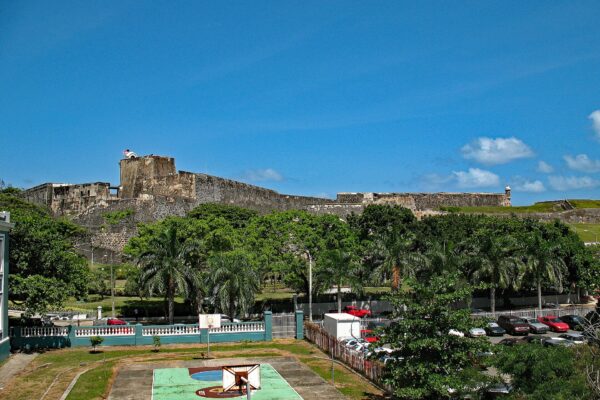 The stone walls and towers of San Cristóbal Fort rise above palm trees and a parking lot in Old San Juan under a deep blue sky, with a neighbourhood basketball court in the foreground