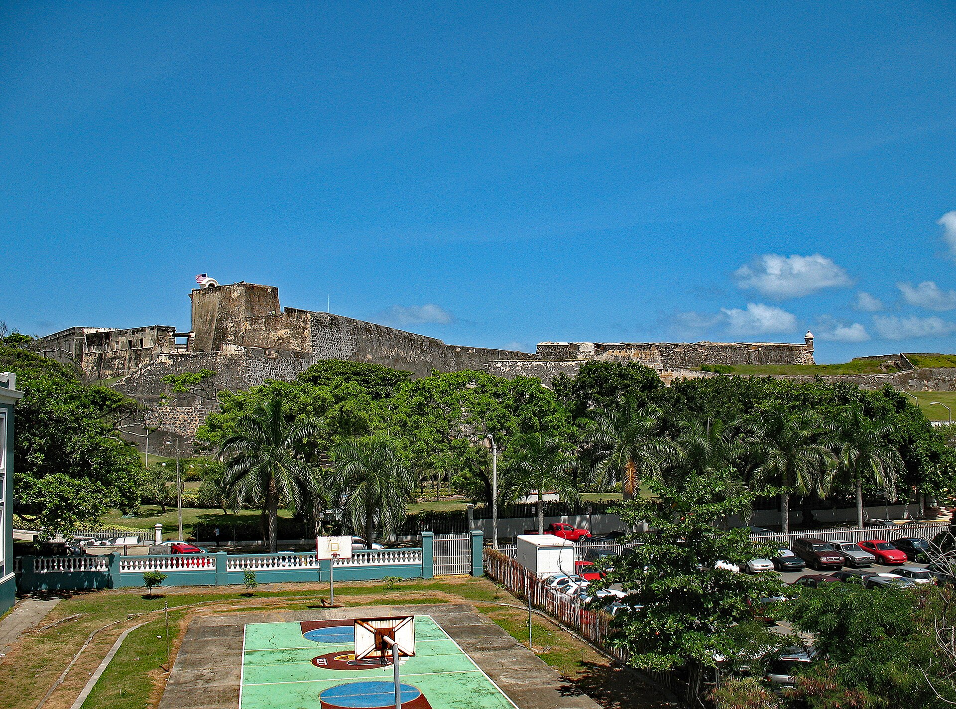 The stone walls and towers of San Cristóbal Fort rise above palm trees and a parking lot in Old San Juan under a deep blue sky, with a neighbourhood basketball court in the foreground