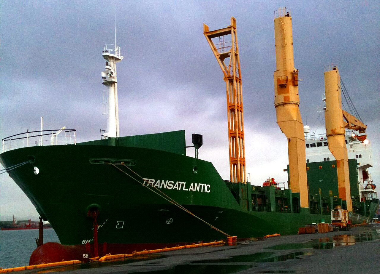 A green cargo ship named TransAtlantic with tall yellow and orange cranes moored at a quayside under a heavy grey sky