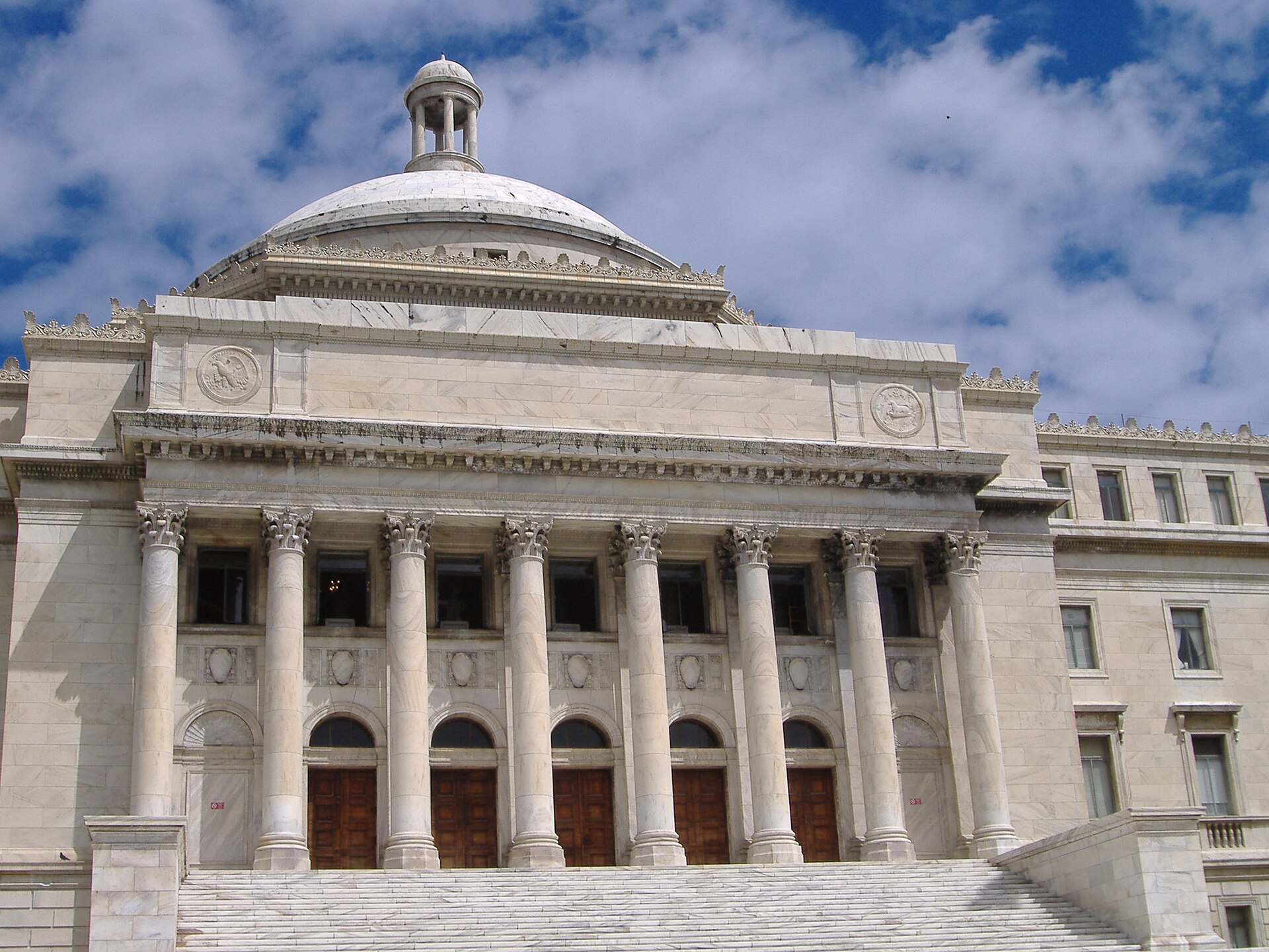 The white marble front of the Capitol of Puerto Rico, with a row of columns, a low dome and wooden doors above a flight of steps, under a blue sky with clouds