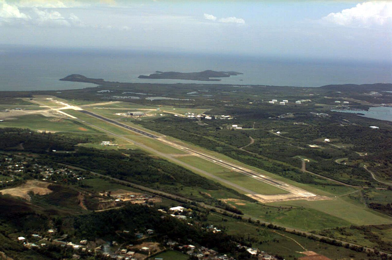Aerial view of a long airfield runway with taxiways and green hills running down to the sea, with two small islands on the horizon