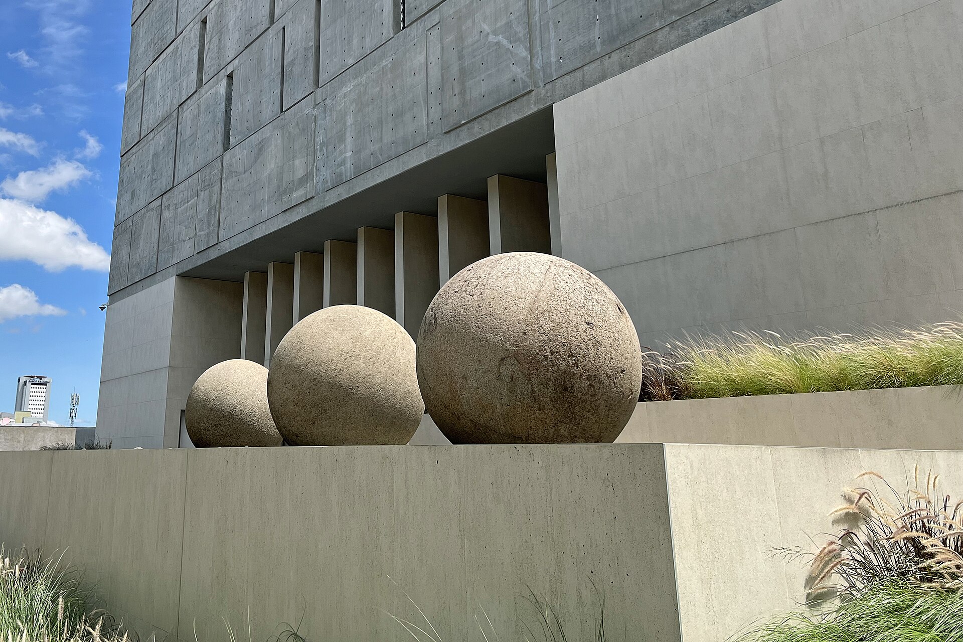 Three large round stone spheres on a concrete ledge in front of a grey concrete building under a blue sky in San José