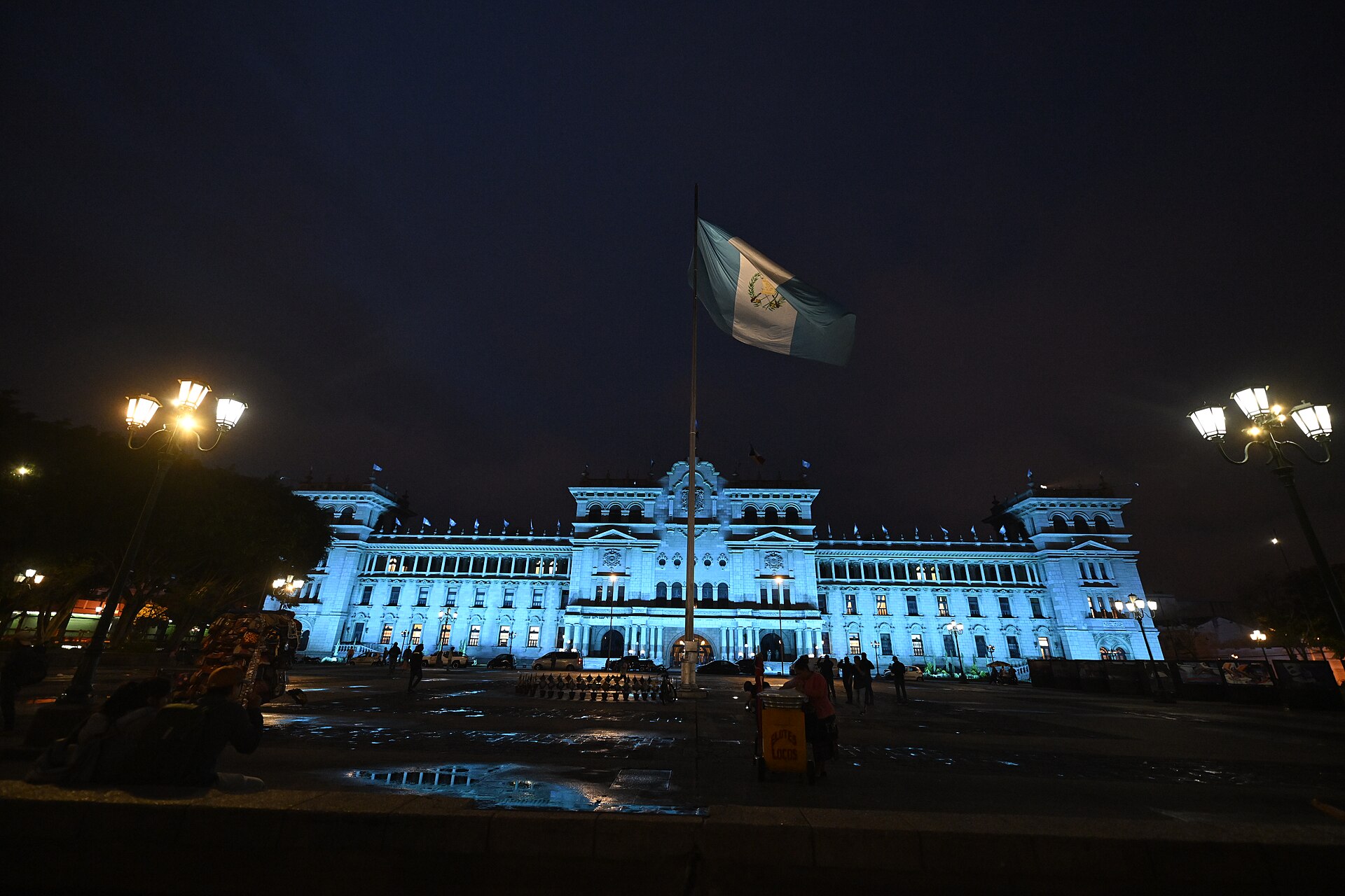 The National Palace of Culture in Guatemala City lit in pale blue at night, with a large Guatemalan flag flying on a pole in the plaza in front and a few people and street lamps around