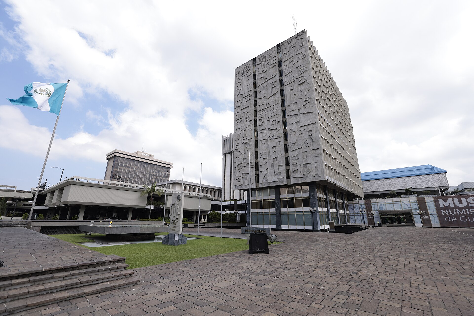 The Bank of Guatemala tower with relief panels on its facade, a Guatemalan flag and a paved plaza in front