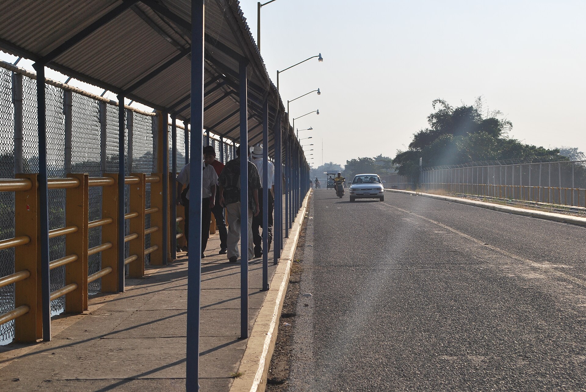 A covered footpath with yellow railings and mesh fencing beside a road on a bridge, with several people walking along it, a car and a motorbike ahead and trees in the haze beyond