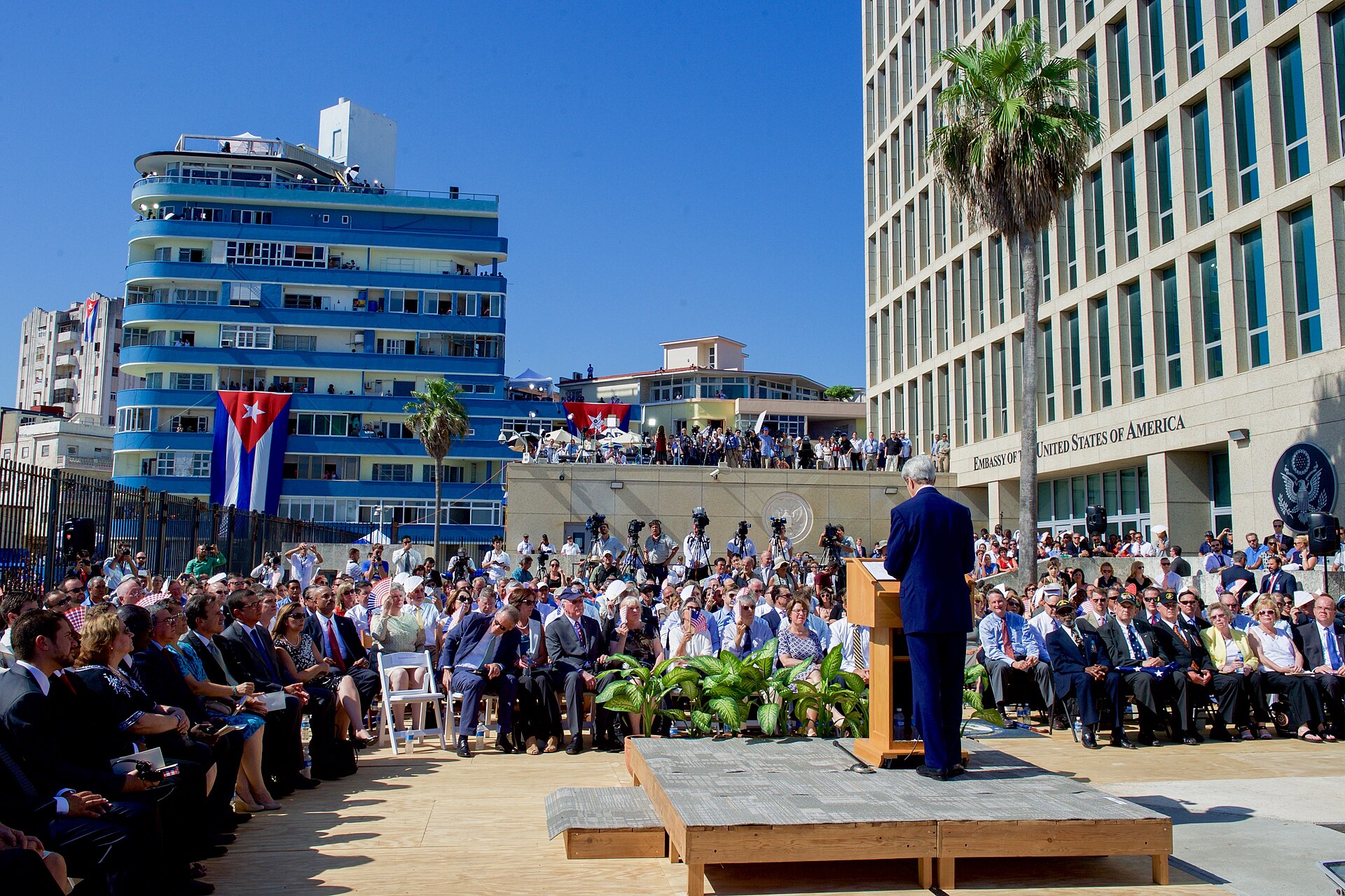 John Kerry speaks at a lectern in front of a seated crowd beside the US Embassy building in Havana, with a Cuban flag on a building behind