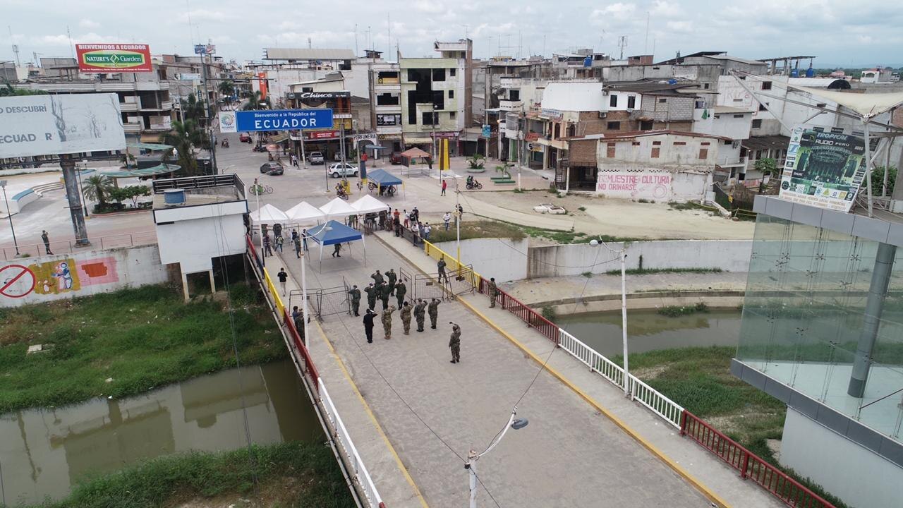 Aerial view of a border bridge with soldiers standing on it, a sign reading Bienvenidos a Ecuador, market tents and buildings beyond and a canal below