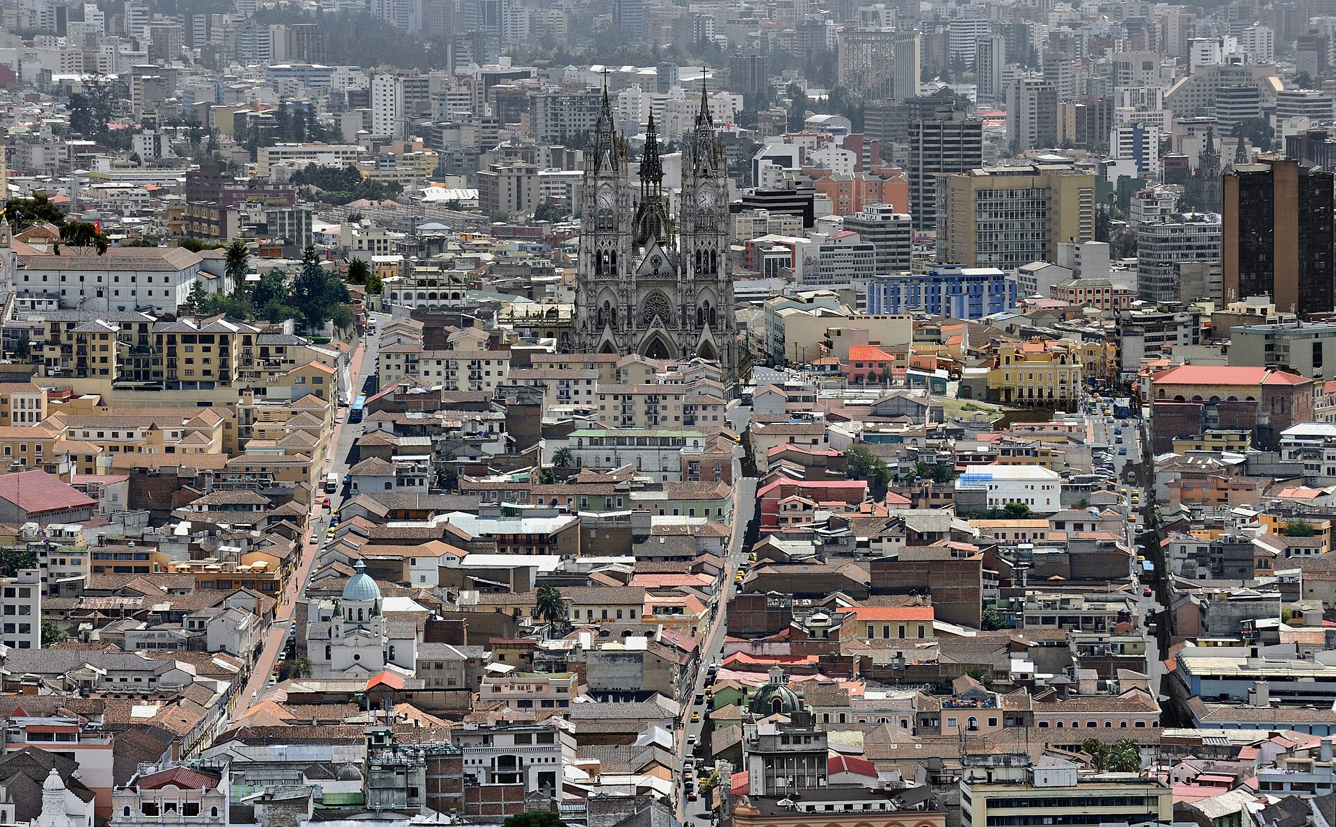 Dense rooftops of Quito's historic centre with a twin-spired neo-Gothic basilica at the centre of the image and modern towers behind