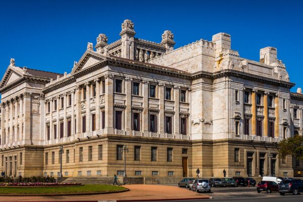 The cream-coloured marble Legislative Palace in Montevideo with its columned wings and carved roofline under a deep blue sky, flower beds and parked cars in front