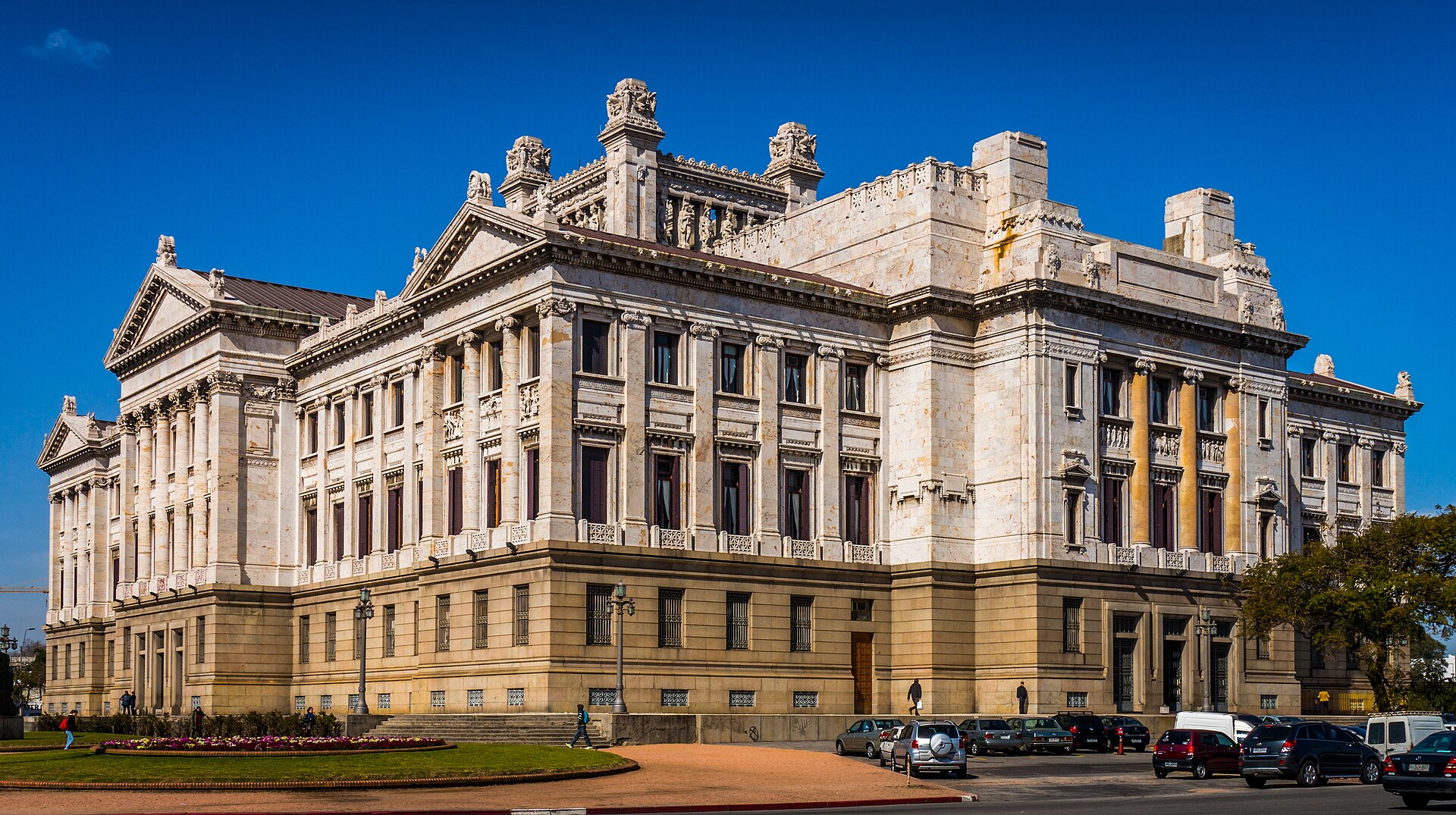 The cream-coloured marble Legislative Palace in Montevideo with its columned wings and carved roofline under a deep blue sky, flower beds and parked cars in front