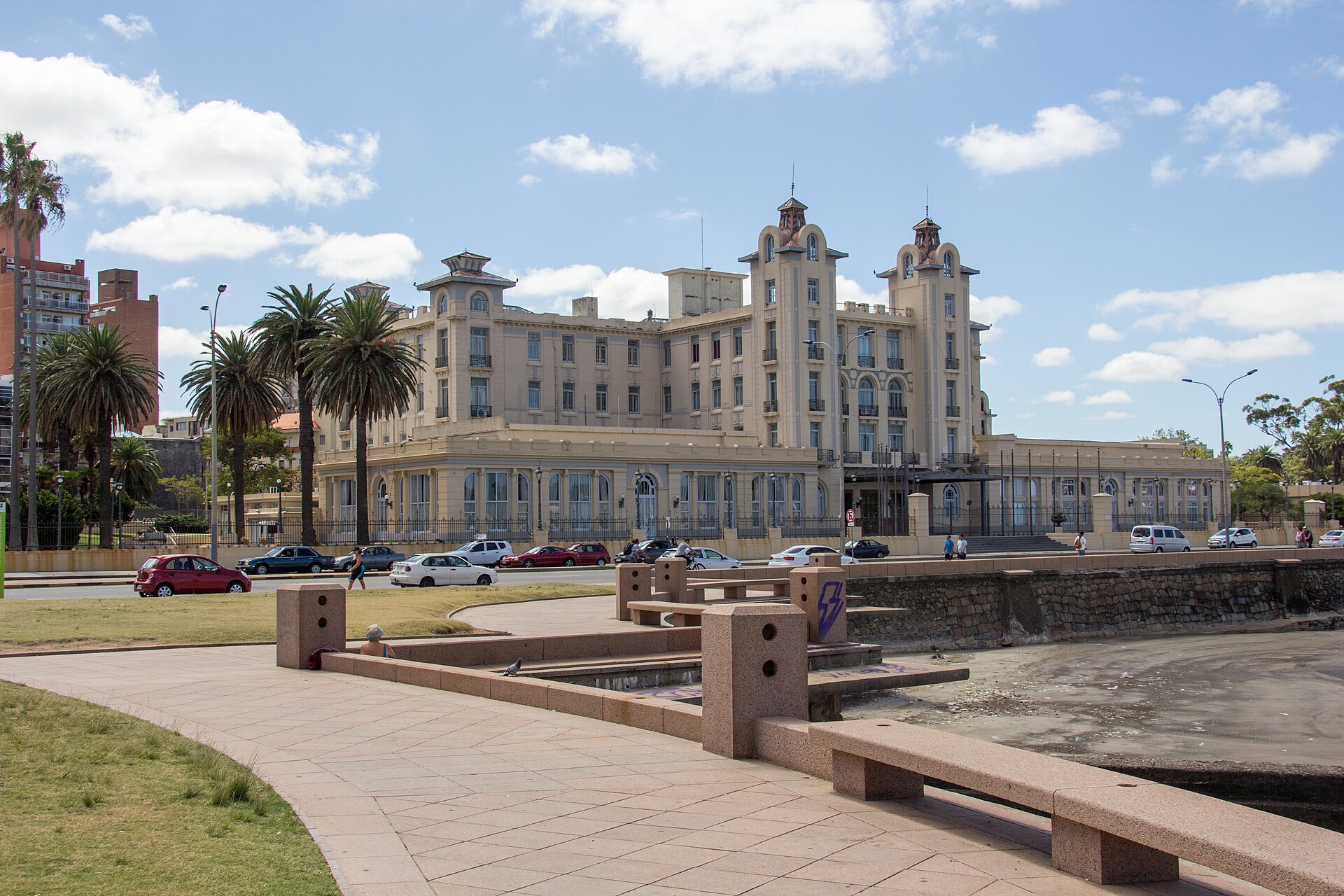 A cream-coloured early twentieth-century building with two corner towers, palm trees and a fence, seen from a paved seafront walkway with granite benches and a few parked cars