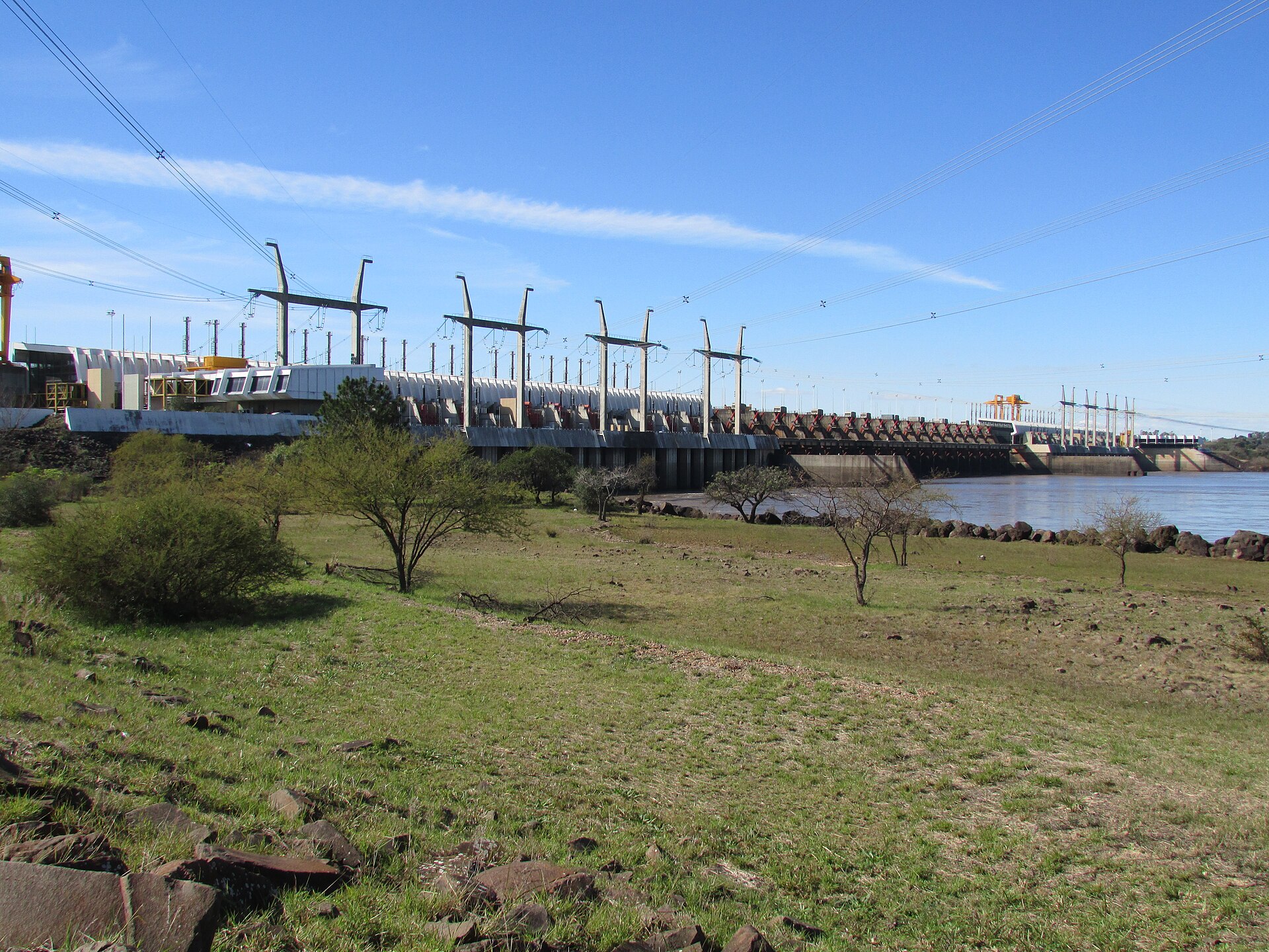The long concrete Salto Grande dam across the Uruguay River with power pylons, a grassy bank with small trees in the foreground and a blue sky with thin clouds