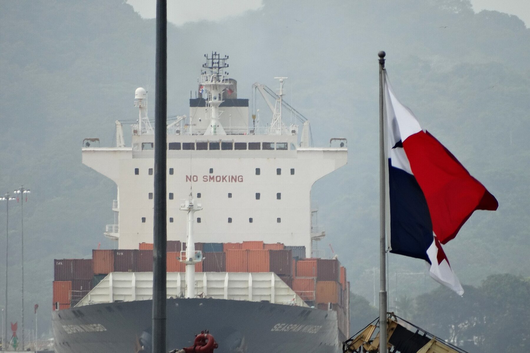 A container ship bow seen head-on at the Miraflores Locks of the Panama Canal, with a Panamanian flag flying in the foreground and green hills behind
