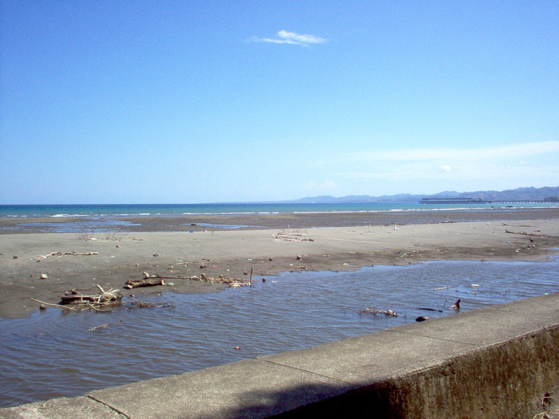 A grey sand beach at Puerto Armuelles in western Panama under a blue sky, with a port pier and the Punta Burica peninsula on the horizon