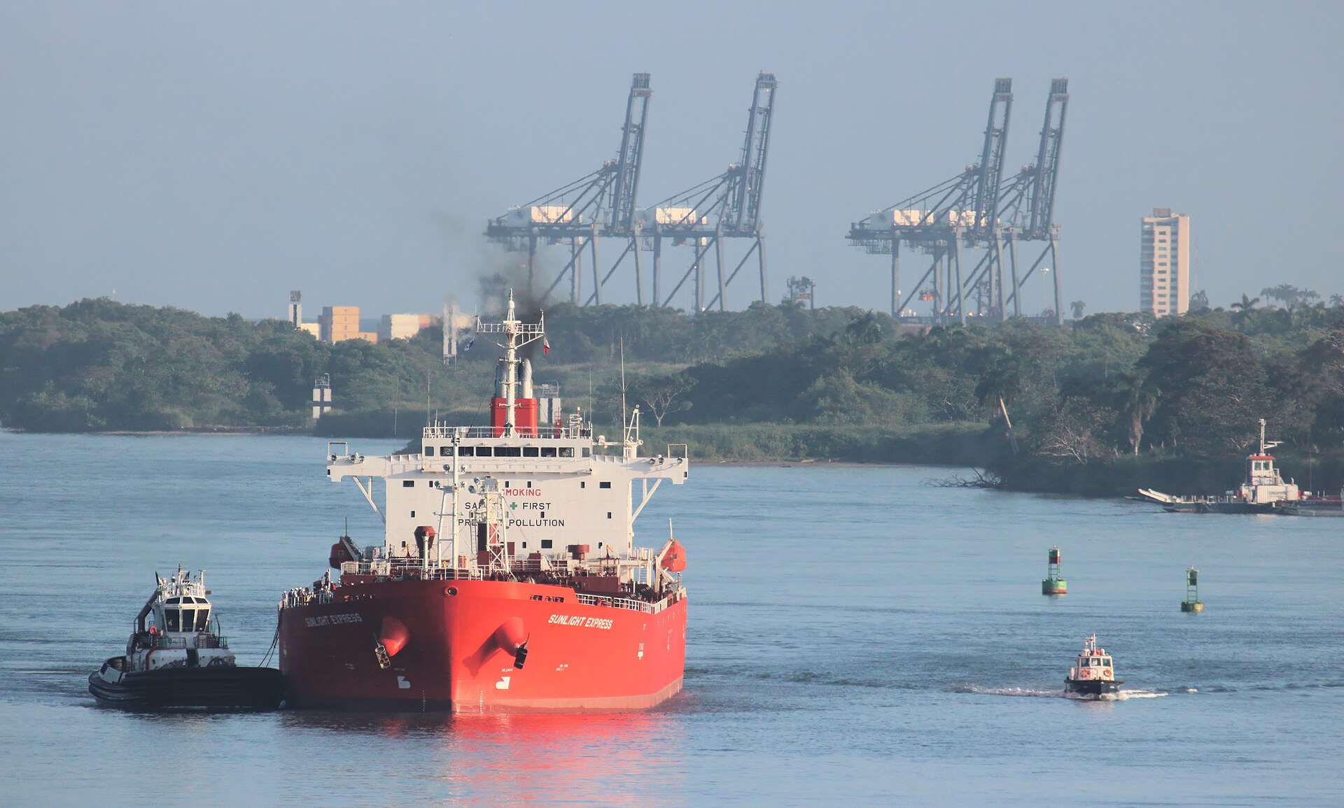 A red oil tanker escorted by tugboats in the Panama Canal's Caribbean entrance near Colón, with container cranes behind