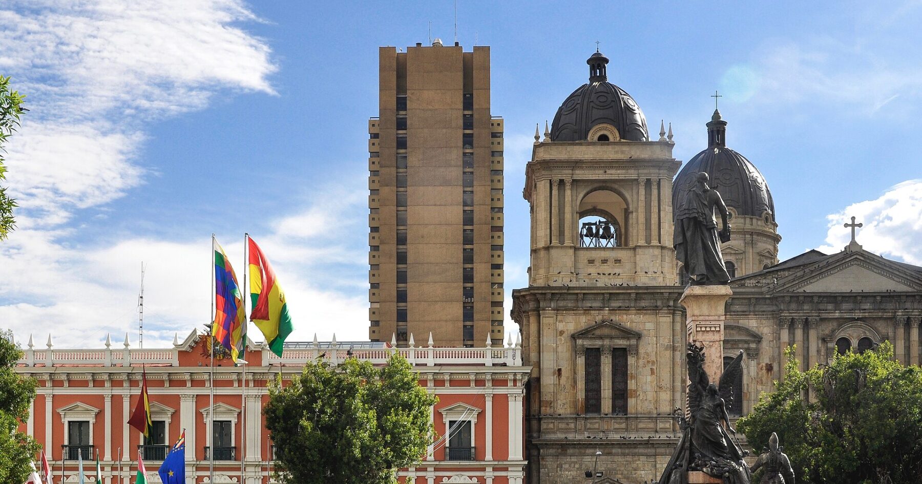 A red and white colonial palace with the Bolivian and Wiphala flags stands beside a domed stone cathedral, with a tall brown tower behind, on a sunny square in La Paz