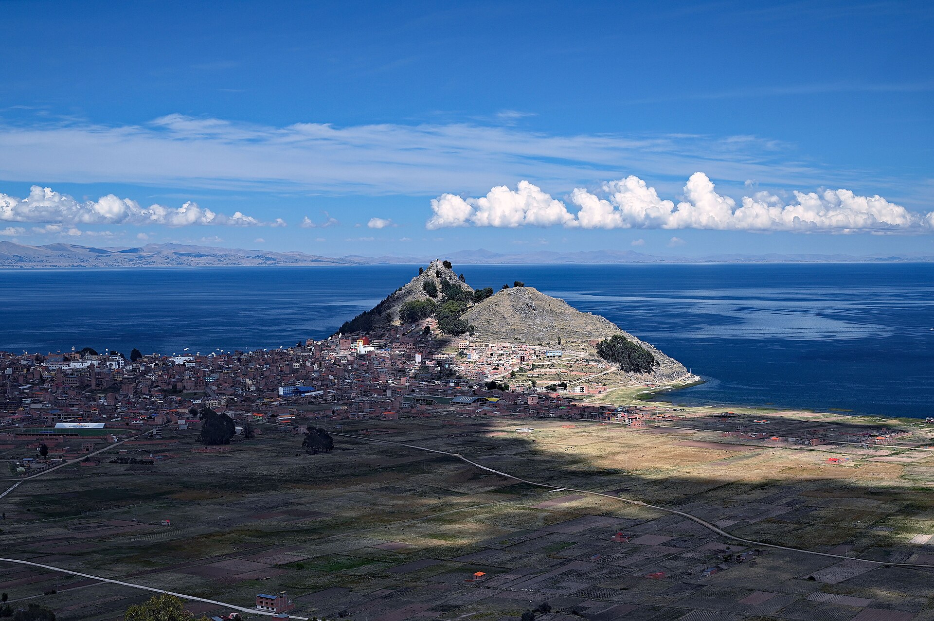 A town of brown rooftops on a hillside peninsula beside the deep blue water of Lake Titicaca, with farmland in the foreground and clouds above