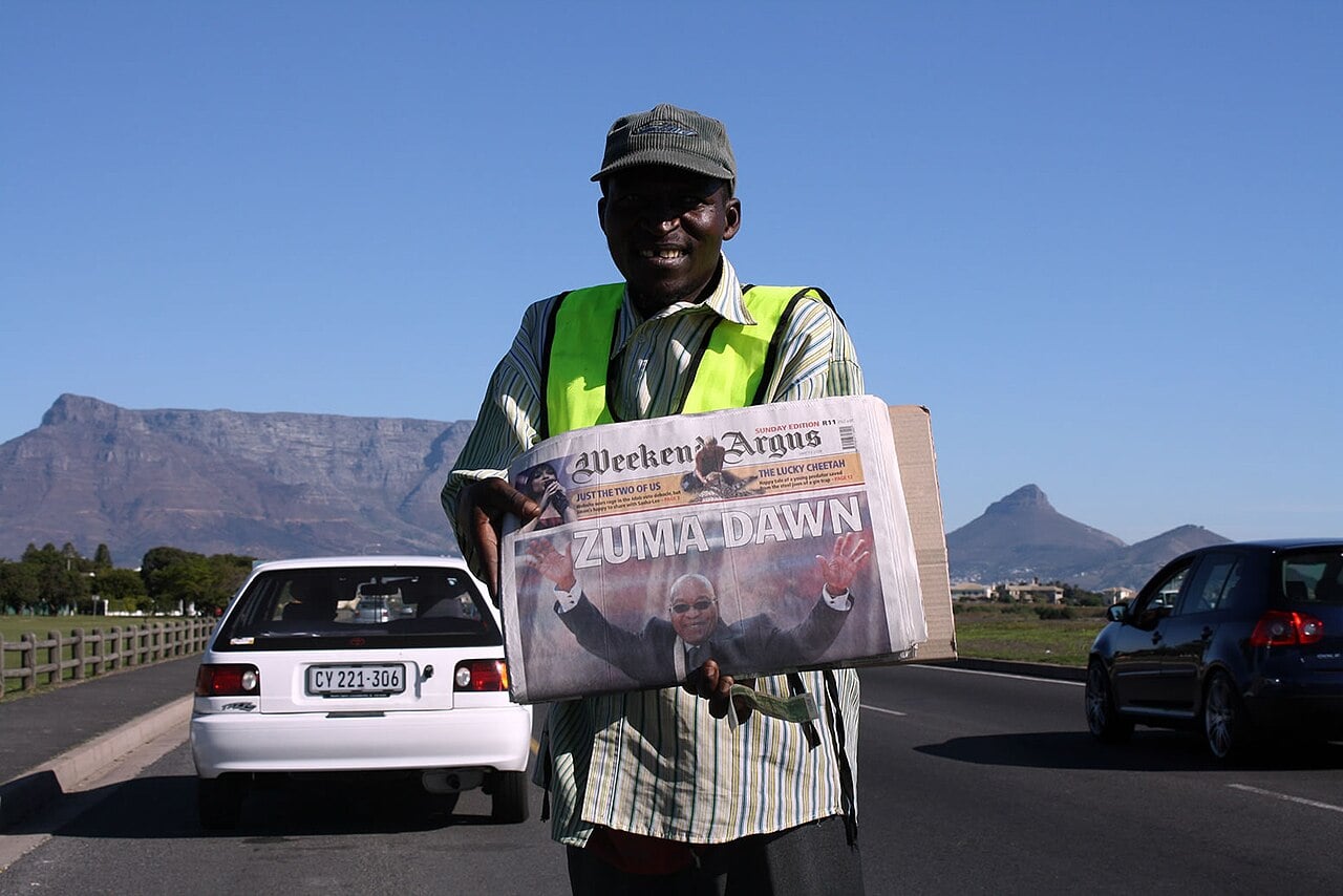 A smiling newspaper seller in a hi-vis vest holds up a Weekend Argus front page reading Zuma Dawn on a Cape Town road, with Table Mountain behind him
