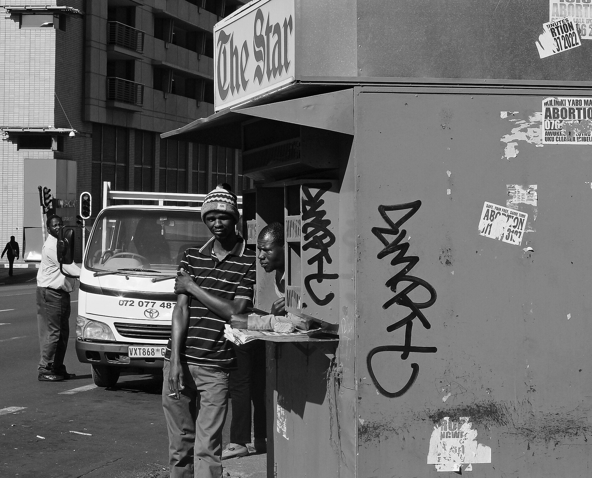 A black-and-white street scene with a newspaper kiosk under a The Star sign, a man in a striped shirt beside it, a white delivery van and a traffic light behind