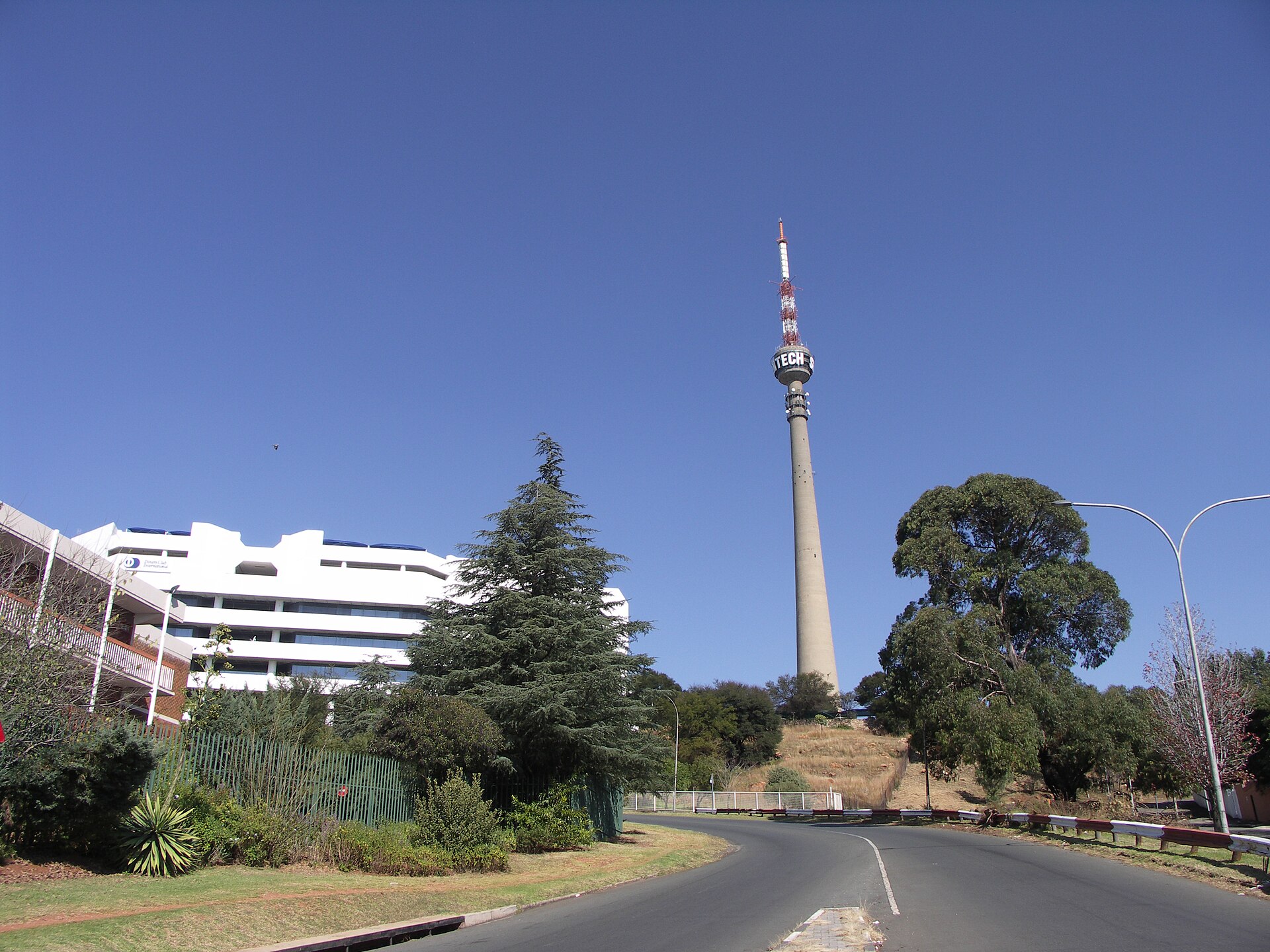A tall concrete tower with a red-and-white mast and a partly visible Sentech sign stands on a hill above a curving road, trees and a white office building under a clear blue sky