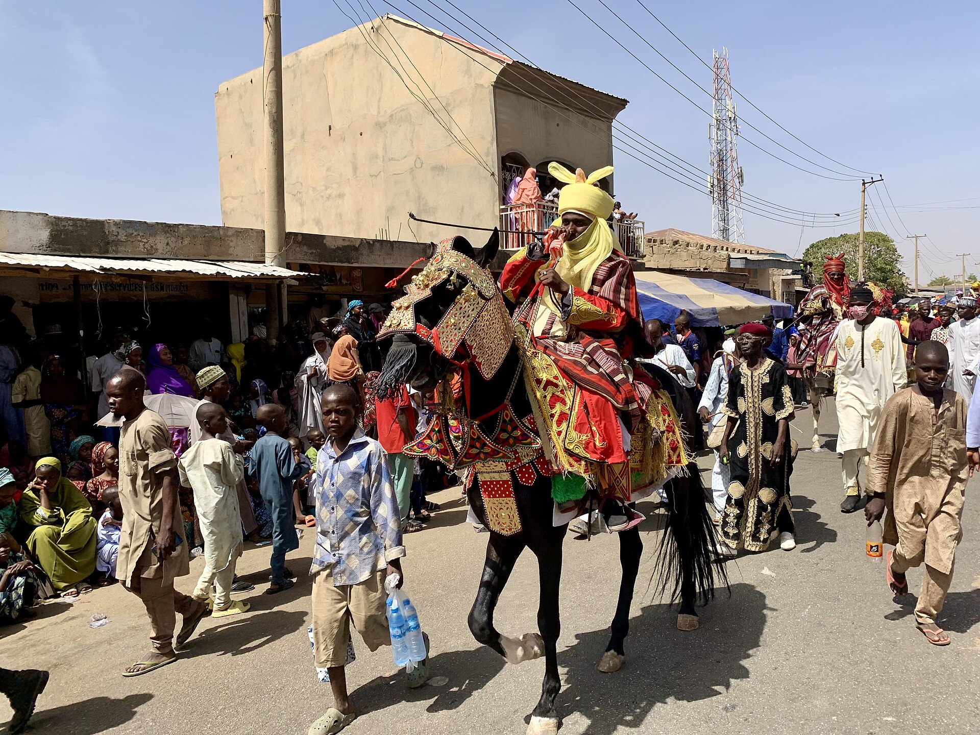 A horseman in a yellow turban and red embroidered robe rides a decorated black horse past a crowd in a street in Katsina