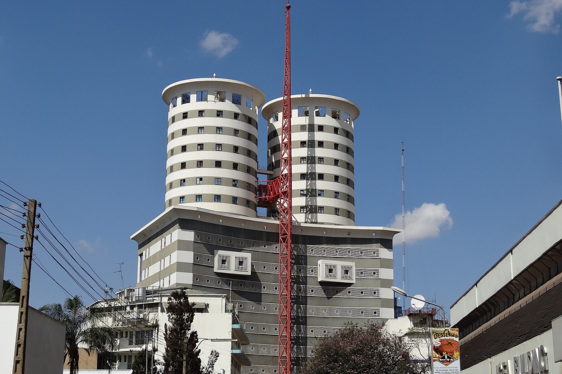 Two tall cylindrical striped office towers with a red mast in front of them rise above low buildings under a blue sky in central Nairobi
