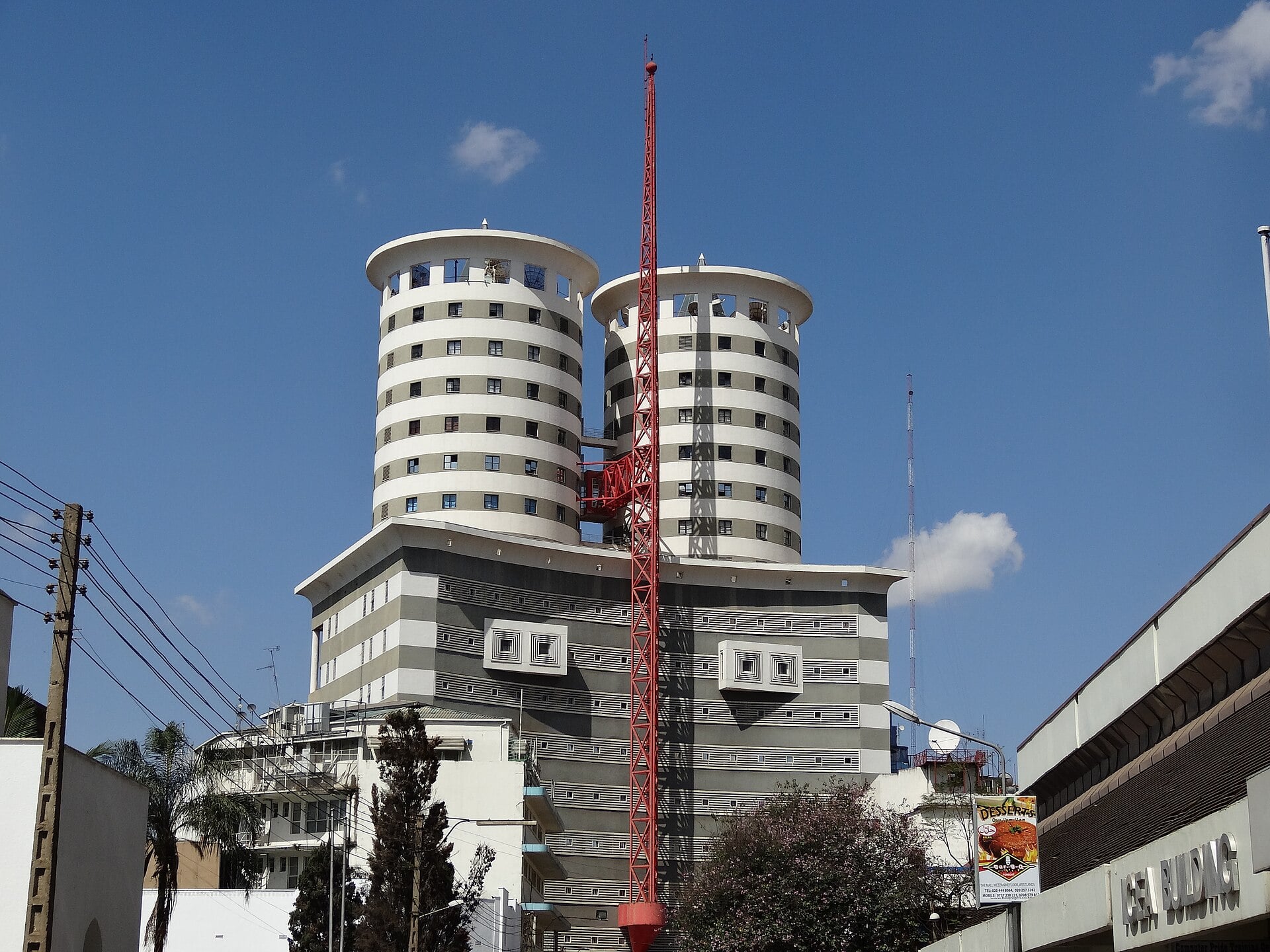 Two tall cylindrical striped office towers with a red mast in front of them rise above low buildings under a blue sky in central Nairobi