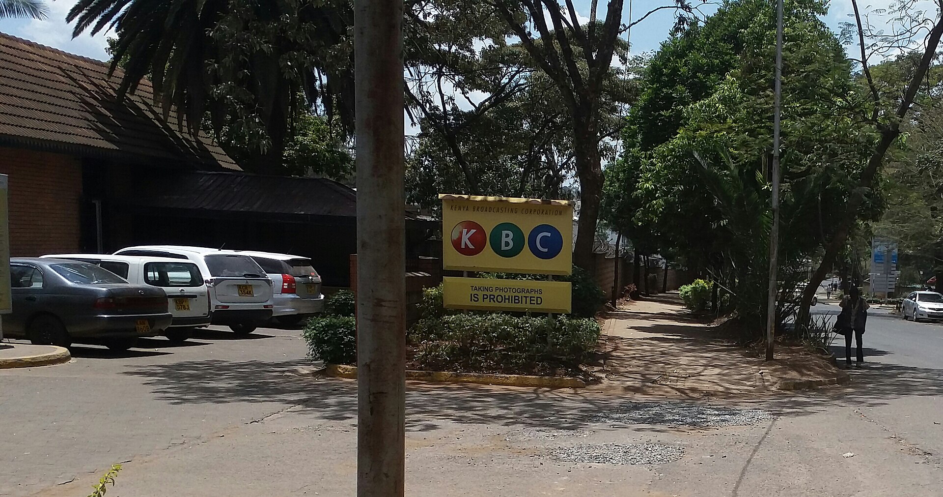 A yellow sign with the red, green and blue KBC logo stands among shrubs beside a car park and a tree-lined path in Nairobi